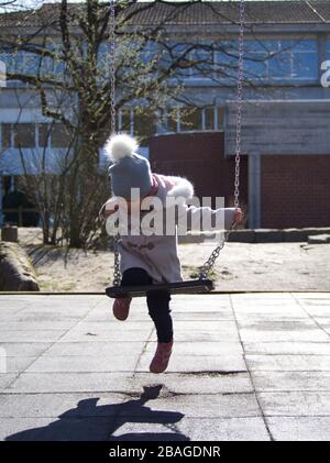 Cute little blonde caucasian girl in winter clothes using a swing at a children play ground in bright sun light Stock Photo