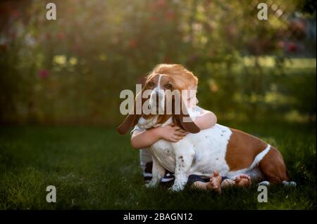 Toddler boy sitting and hugging his hound dog in the backyard grass Stock Photo