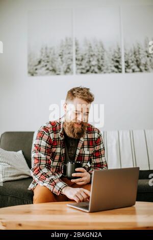 man with beard holds cup of coffee while working on laptop computer Stock Photo
