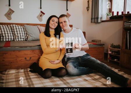 Pregnant woman and boyfriend with baby scan.Young couple expecting baby sitting on bed holding medical baby scans. Stock Photo
