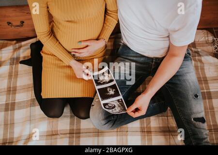 Young couple holding baby scan photos on bed. High angle of young man and woman expecting baby sitting on bed holding baby scans. Stock Photo