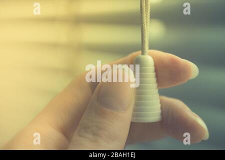 rope blinds with plastic cap in female fingers close-up, background horizontal shutters. Stock Photo