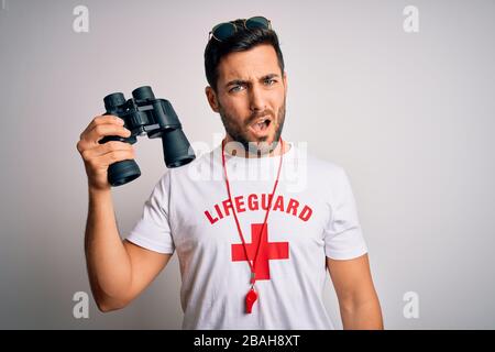 Young lifeguard man with beard wearing t-shirt with red cross and sunglasses using whistle In shock face, looking skeptical and sarcastic, surprised w Stock Photo