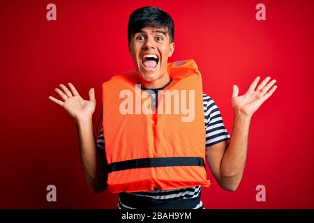 Young handsome man wearing orange safety life jacket over isolated red background celebrating crazy and amazed for success with arms raised and open e Stock Photo