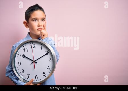 Young little boy kid holding big minute clock over isolated pink background serious face thinking about question, very confused idea Stock Photo