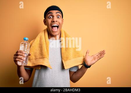 Young handsome african american sportsman wearing towel drinking bottle of water very happy and excited, winner expression celebrating victory screami Stock Photo