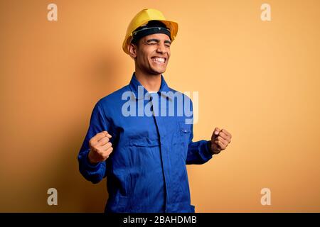Young handsome african american worker man wearing blue uniform and security helmet very happy and excited doing winner gesture with arms raised, smil Stock Photo