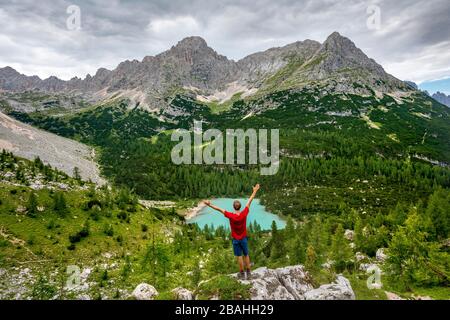 Young man stretches arms in the air, hiker stands on rocks and looks at turquoise green Sorapiss lake and mountain landscape, Dolomites, Belluno Stock Photo