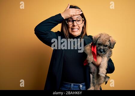 Young beautiful brunette woman holding cute puppy pet over isolated yellow background stressed with hand on head, shocked with shame and surprise face Stock Photo