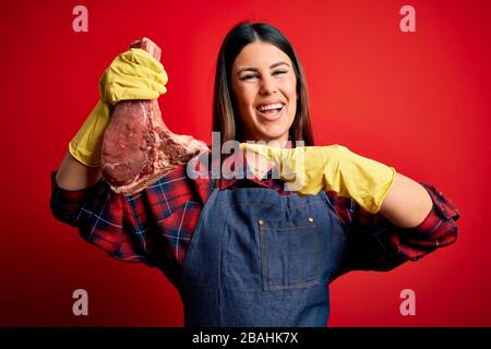 Young butcher woman holding fresh raw beef meat stake over red background very happy pointing with hand and finger Stock Photo