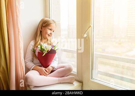 School kid in self isolation sitting near window with bouquet of flowers. Girl stay at home during covid-19 lock down, self isolation, stay home Stock Photo