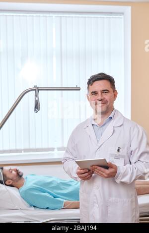 Portrait of smiling senior doctor in lab coat standing with tablet in patients room Stock Photo
