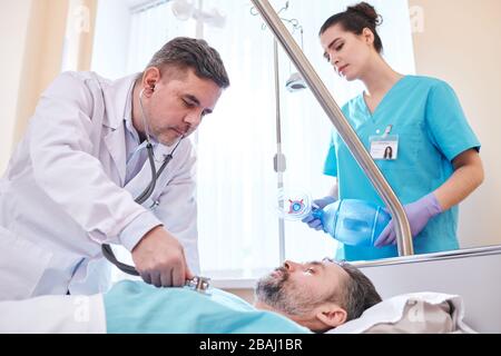 Concentrated mature doctor using stethoscope while examining lungs of patient in hospital room, nurse with ambu mask standing behind bed Stock Photo