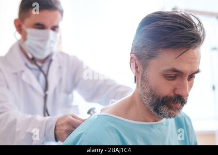 Doctor in surgical mask using stethoscope while listening to patients lungs with stethoscope Stock Photo