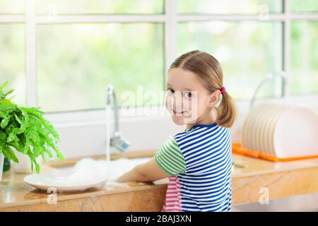 Child washing dishes. Home chores. Kid in white kitchen cleaning plates after lunch at window. Stock Photo