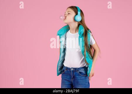 A little girl with headphones explodes pink chewing gum on a pink background. Stock Photo