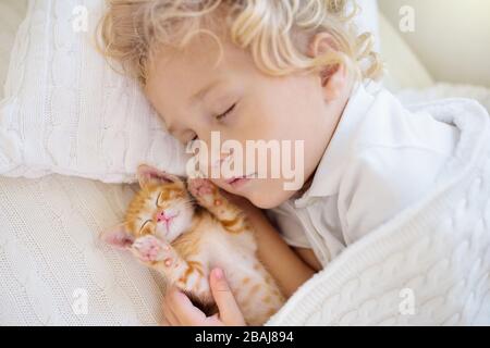 Baby boy sleeping with kitten on white knitted blanket. Child and cat. Kids and pets. Little kid with his animal. Cozy winter evening with pet. Childr Stock Photo