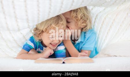 Child reading book in bed under knitted blanket. Two brothers playing together. Kids cozy bedroom hygge style. Little boy doing homework before sleep. Stock Photo