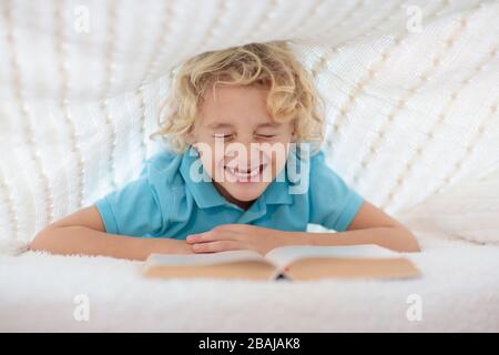 Child reading book in bed under knitted blanket. Kids cozy bedroom hygge style. Little boy doing homework before sleep. Stock Photo