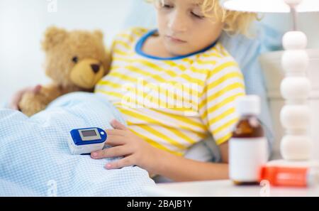 Sick little boy with pulse oximeter on his finger. Asthma treatment. Ill child lying in bed. Unwell kid with chamber inhaler, cough medicine. Flu seas Stock Photo