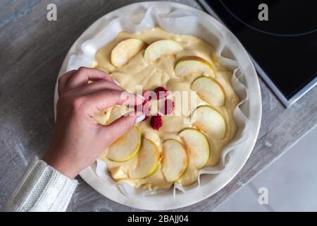 The hostess at home is preparing apple charlotte in the kitchen. puts the berry on an apple pie in a baking dish. Raw Apple Pie Dough Stock Photo
