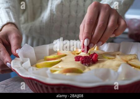 The hostess at home is preparing apple charlotte in the kitchen. puts the berry on an apple pie in a baking dish. Raw Apple Pie Dough Stock Photo