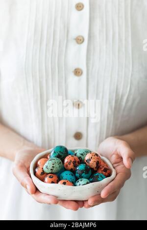 Woman hands holding small beautiful ceramic plate with painted quail eggs for Easter. White rustic dress, red and blue trendy color of eggs. Stock Photo