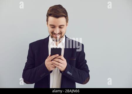 Positive guy in a jacket is typing text in a mobile smiling with on a gray background. Stock Photo