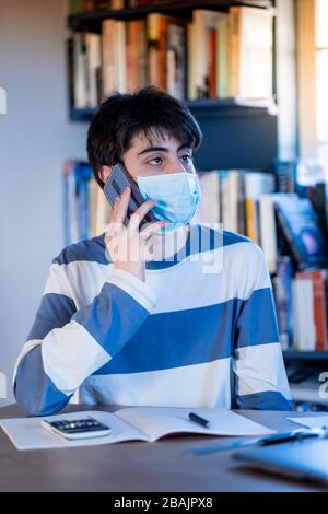 A teenager wearing a face mask, sitting in front of their homework and calling their friends on their mobile Stock Photo