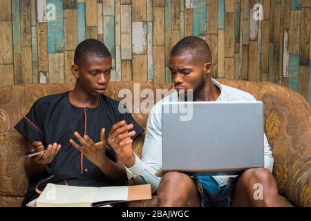 two african young men sitting down discussing about their work also holding a laptop to work online Stock Photo