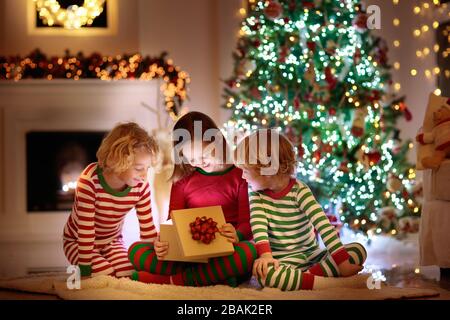 Children at Christmas tree and fireplace on Xmas eve. Family with kids celebrating Christmas at home. Boy and girl in matching pajamas decorating xmas Stock Photo