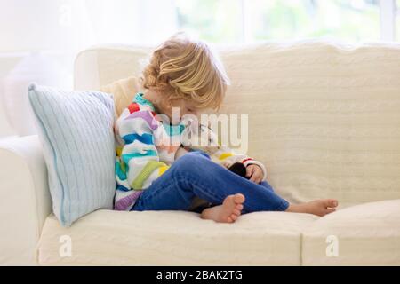 Child playing with cat. Kid holding kitten. Little boy snuggling cute pet animal sitting on couch in sunny living room at home. Kids play with pets. C Stock Photo