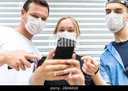 Three young people of different ethnicities with protective masks on their faces using a mobile phone on the street. Covid-19 Stock Photo