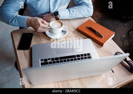 Young serious businessman working on computer at table, drinking coffee. Man analyzes information, data, checking email. Freelancer working at home. O Stock Photo