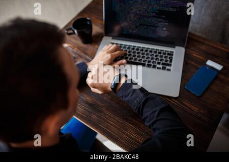 a male programmer in a black shirt is working at a laptop, close-up of the program code and developer's hands on a laptop. modern young programmer sit Stock Photo