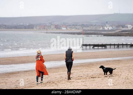 Portobello, Scotland, UK. 28 March, 2020. On the first weekend of the coronavirus lockdown the public were outdoors exercising and maintaining social distancing along Portobello beachfront promenade. Two females walking dog on beach.  Iain Masterton/Alamy Live News Stock Photo