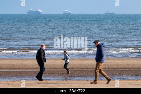 Portobello, Scotland, UK. 28 March, 2020. On the first weekend of the coronavirus lockdown the public were outdoors exercising and maintaining social distancing along Portobello beachfront promenade. Pictured People walking and jogging on beach with three out of service cruise liners from Fred Olsen at anchor in distance. Iain Masterton/Alamy Live News Stock Photo