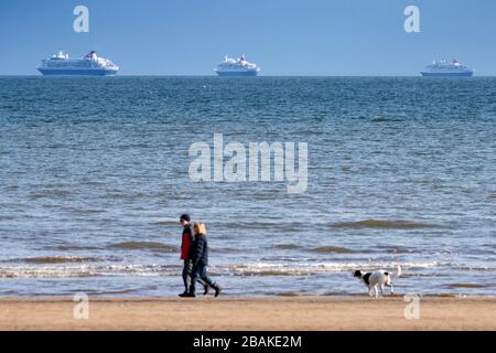 Portobello, Scotland, UK. 28 March, 2020. On the first weekend of the coronavirus lockdown the public were outdoors exercising and maintaining social distancing along Portobello beachfront promenade. Pictured People walking dog on beach with three out of service cruise liners from Fred Olsen at anchor in distance. Iain Masterton/Alamy Live News Stock Photo