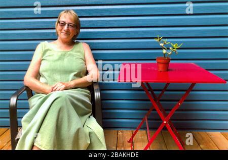 A happy, smiling, contented woman is relaxing on her house deck. Wearing a pale green summer dress, she sits with her hands folded in her lap and her legs crossed.  A bright red, scissor-legged table stands beside her, in front of a wall of bright blue, wood-grained vinyl siding. A small green succulent Jade plant in a brown pot rests in the center of the red table. Along with the warm brown floorboards, the bright colors and composition create a wonderfully friendly and comforting photograph.   To see my other images, which are mostly vintage, Search:  Prestor  vintage  woman Stock Photo