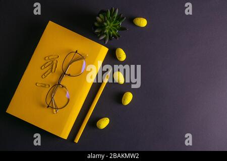 Yellow pencil, yellow notebook glasses, pins, clip paper and yellow easter eggs on on black background Stock Photo