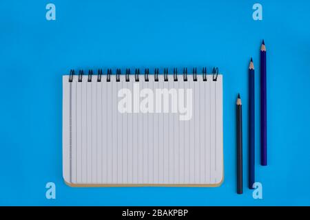 An opened white paper diary beside three wood pencil crayons over a blue paper background Stock Photo