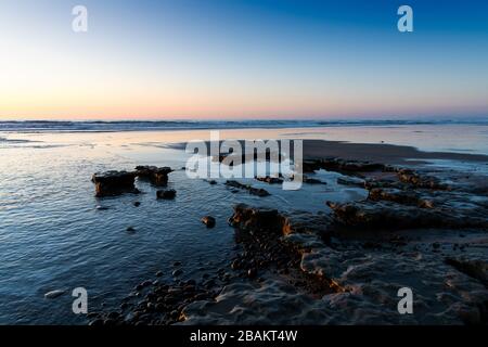 The light of a sunset casts golden highlights in pebbles and low rock formations on a beach under a purple and blue sky - Moonlight Beach, Encinitas, Stock Photo