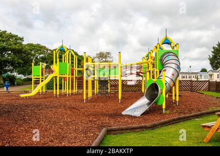 Bright yellow poles support a a variety of children's playground equipment i.e. a tubular slide, swings,  frames, on a brown wood-chip safety surface Stock Photo