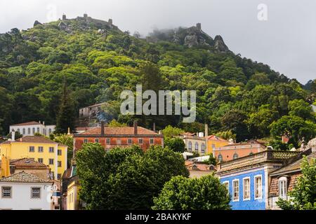 Beautiful, colorful buildings in the village of Sintra, Portugal on a sunny, forested hillside below misty ancient castle ruins Stock Photo