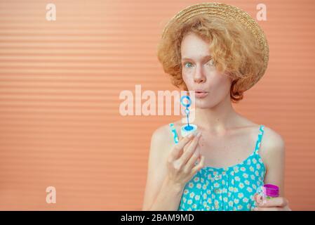 young cheerful curly redhead woman in blue sundress and straw hat with bubbles on beige background. Fun, summer, fashion, youth concept Stock Photo