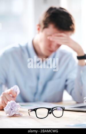 Tired man being puzzled with paperwork sitting at desk with crumpled paper in hand, focus on eyeglasses Stock Photo