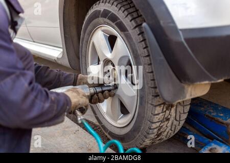 Hands of automotive mechanic unscrewing nuts with pneumatic impact wrench during car wheels season changing. Close-up view with selective focus and ba Stock Photo