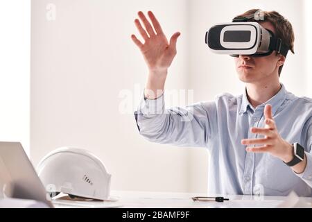 Concentrated young engineer sitting at table and twisting hands while using virtual reality device to make 3D building project Stock Photo