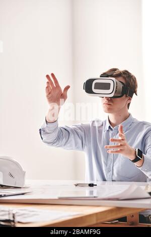 Male engineer sitting at desk and gesturing hands while designing 3D model of building with VR device Stock Photo