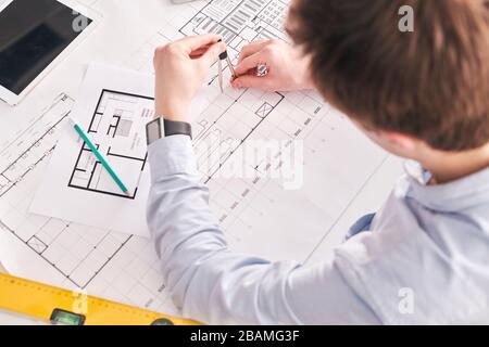 Above view of building engineer sitting at desk and using pair of compasses while working on floor plan of flat Stock Photo